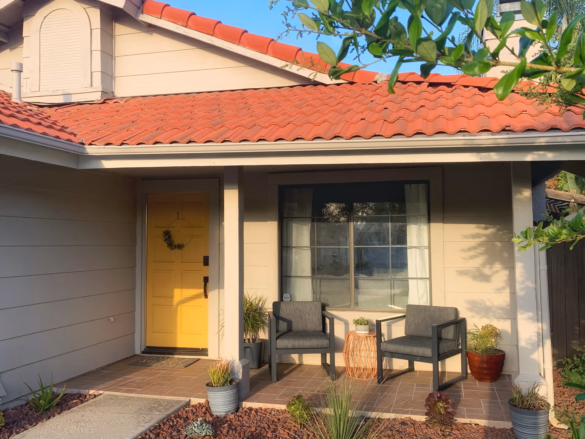 Front porch of a house with a yellow door decorated with a small wreath, two gray cushioned chairs, a small round table with a plant, and several potted plants around the entrance. The house has beige siding and a red tiled roof.