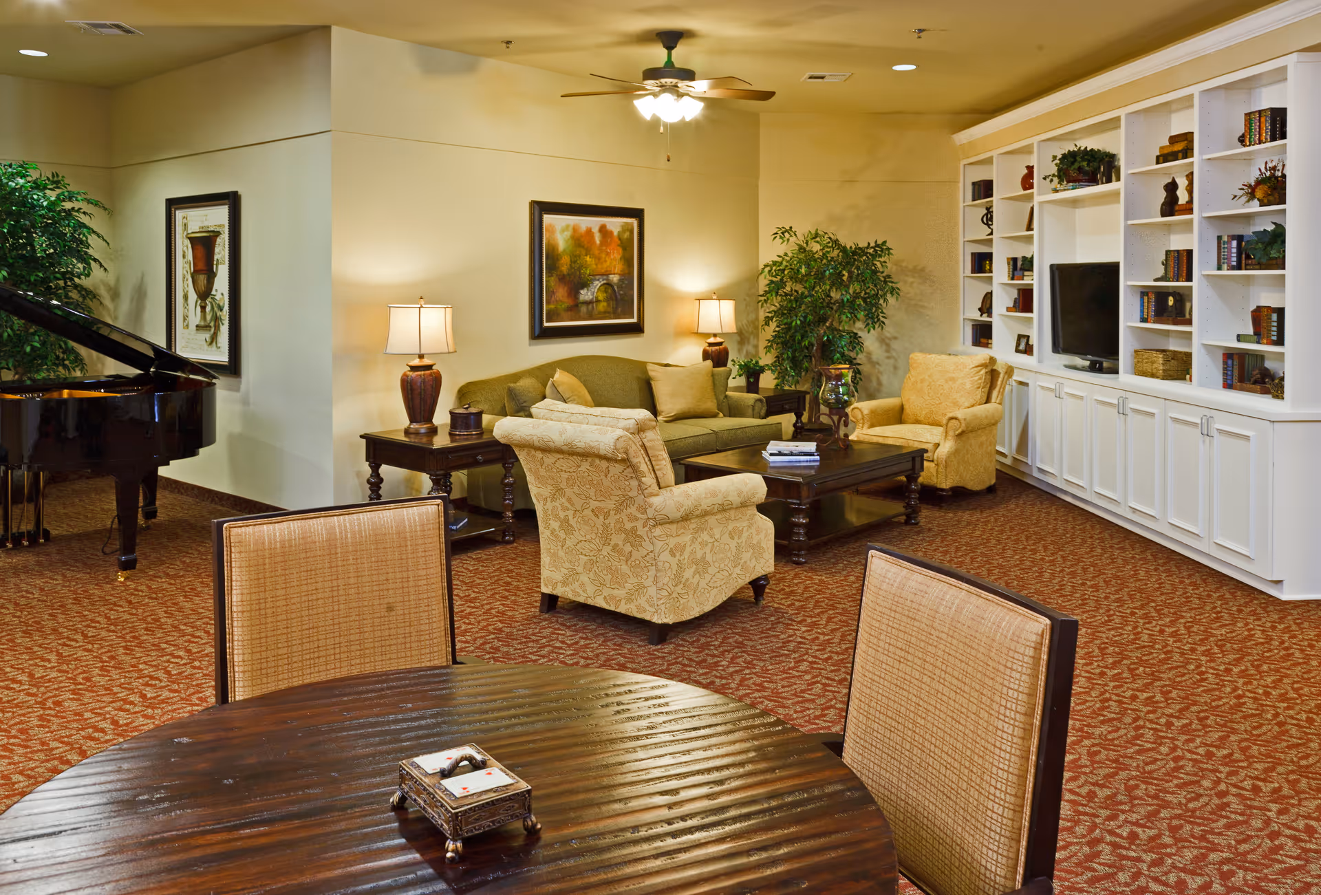 A furnished common lounge with sofas and armchairs around a coffee table, built-in bookshelves, a grand piano, and a dining table in the foreground.