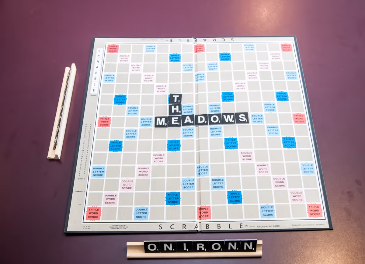 A Scrabble board on a maroon table with the word 'THE MEADOWS' spelled out horizontally in the center. A wooden tile holder below the board displays the letters 'O N I R O N N'. A Scrabble tile rack with additional tiles is placed to the left of the board.