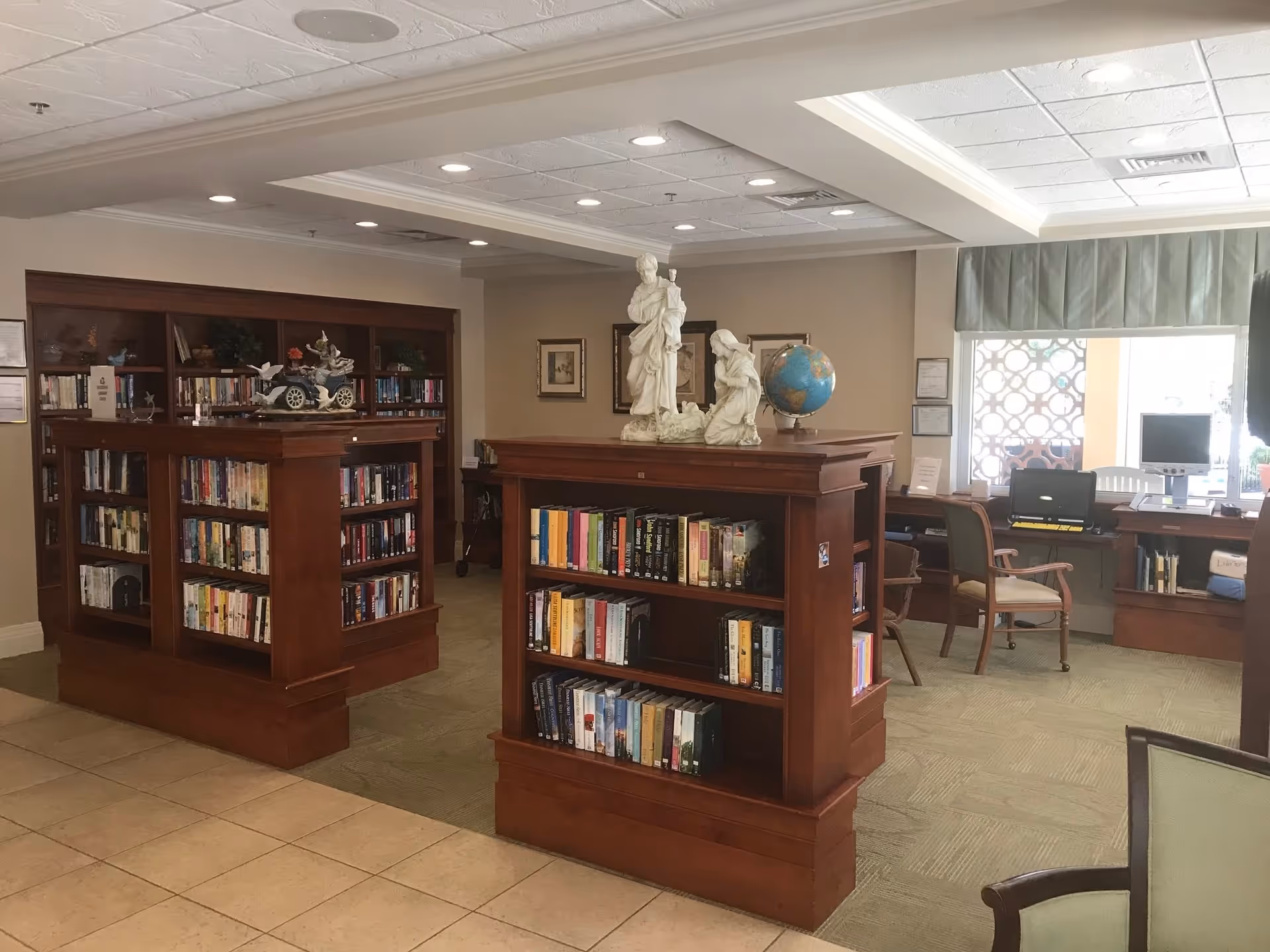 Interior of a library or reading room in a senior living facility with wooden bookshelves filled with books, a globe, and decorative statues on top. There are chairs and computer stations near a window with a patterned screen and green valance.