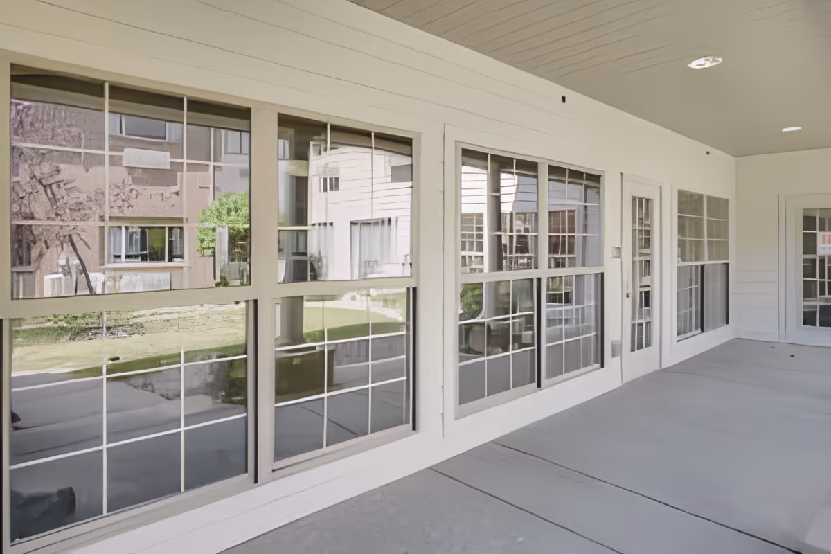 A covered outdoor patio area with large windows and glass doors reflecting the exterior of the building and greenery outside. The patio has a light-colored ceiling with recessed lighting and a smooth concrete floor.