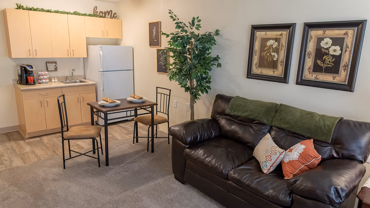 Small living area with a dark leather sofa and pillows, wall art, a kitchenette with fridge and sink, and a small dining table with two chairs.