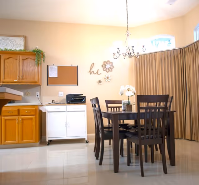 Dining area with a dark wood table and chairs under a chandelier, beige walls, curtains, and kitchen cabinets to the left.