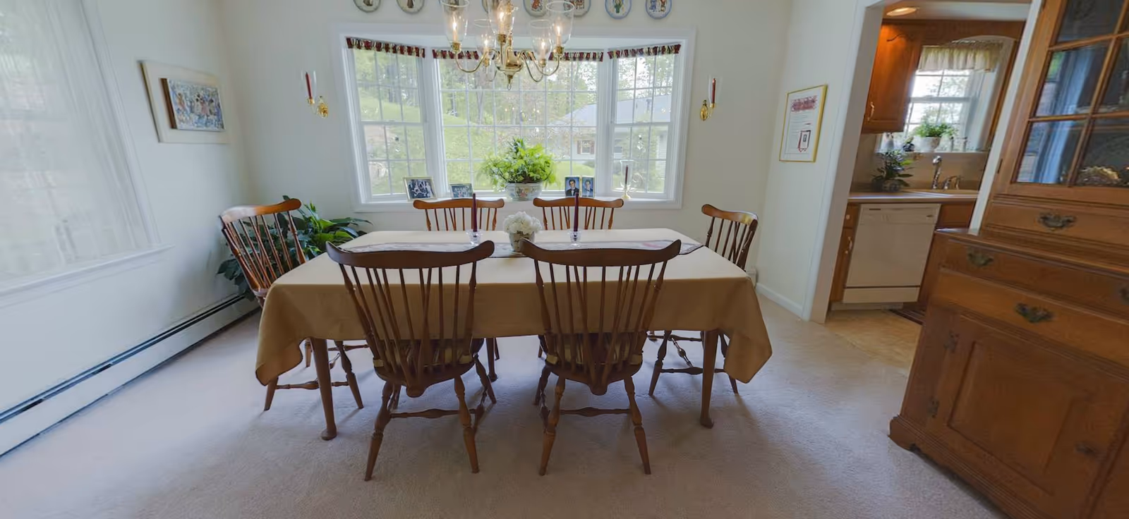 A dining room with a rectangular table covered with a beige tablecloth and six wooden chairs around it. The room has a large bay window with a view of greenery outside. There is a chandelier hanging above the table, and a wooden cabinet is visible on the right side. The adjacent kitchen area can be seen through an open doorway.