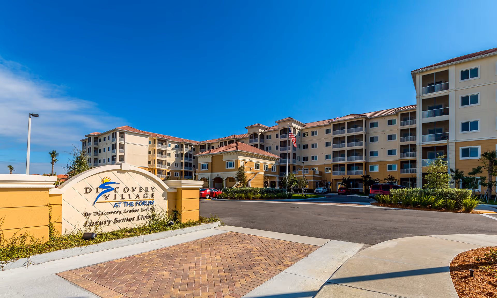 Exterior view of Discovery Village At The Forum senior living facility showing a multi-story building with balconies, a driveway, landscaped greenery, and a sign at the entrance under a clear blue sky.