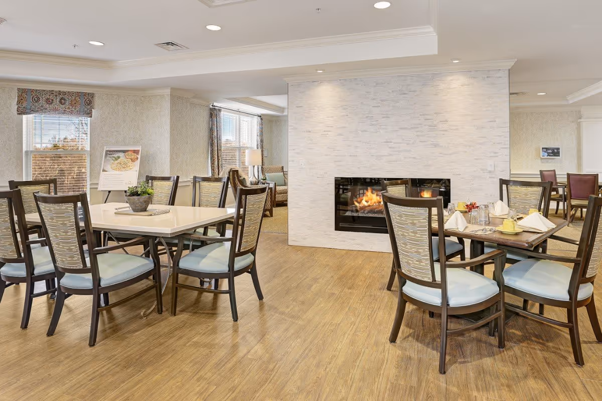Bright dining room with multiple tables and chairs, a modern fireplace set into a white stone wall, and wood-look flooring.