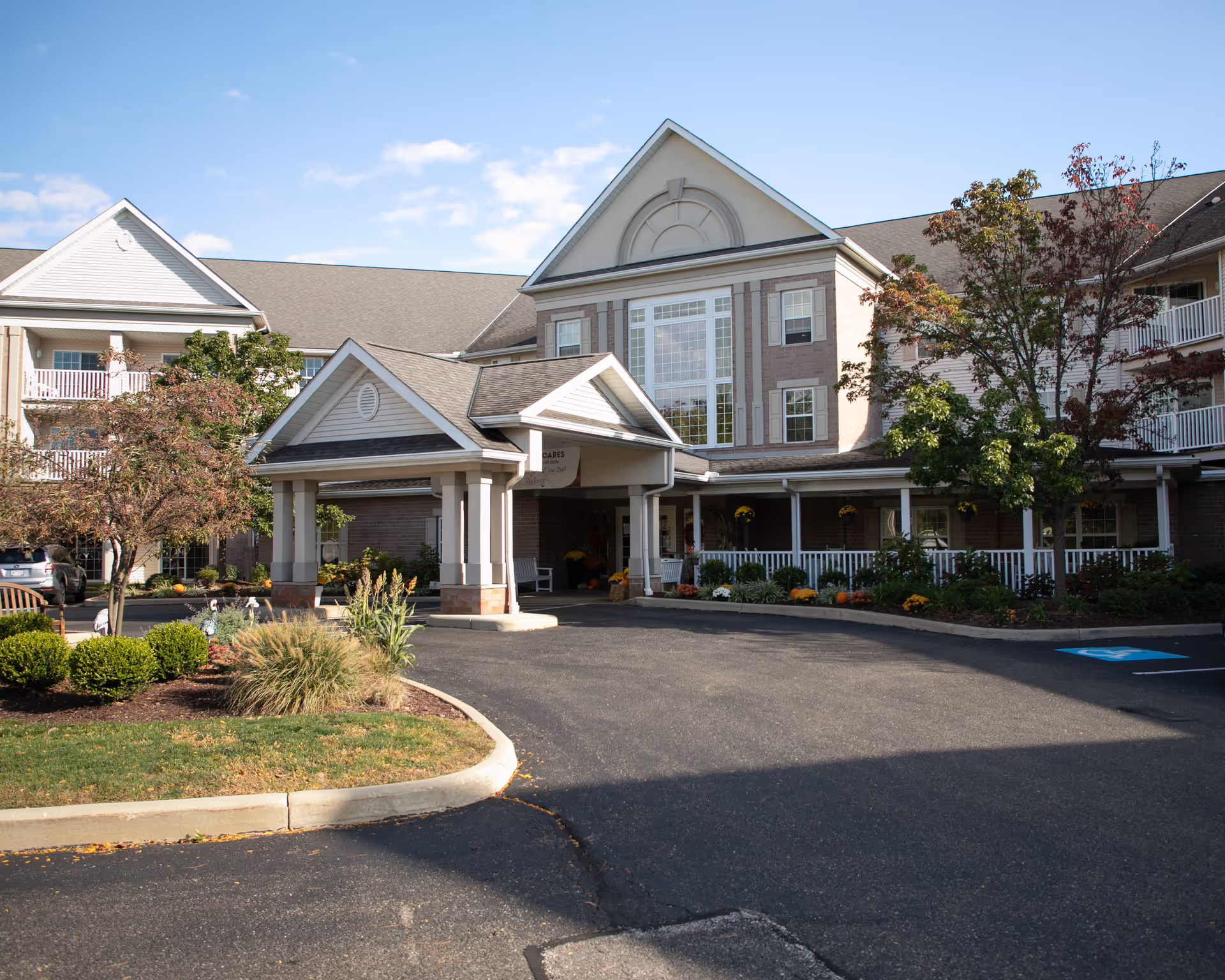 Exterior view of Danbury Cuyahoga Falls senior living facility showing the main entrance with a covered drop-off area, surrounded by landscaped greenery and trees under a partly cloudy sky.
