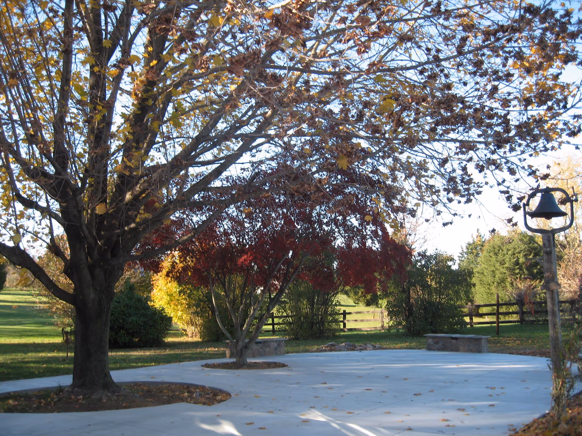 An outdoor area with a concrete pathway surrounded by trees with autumn foliage, a wooden fence in the background, and a metal bell mounted on a post to the right.