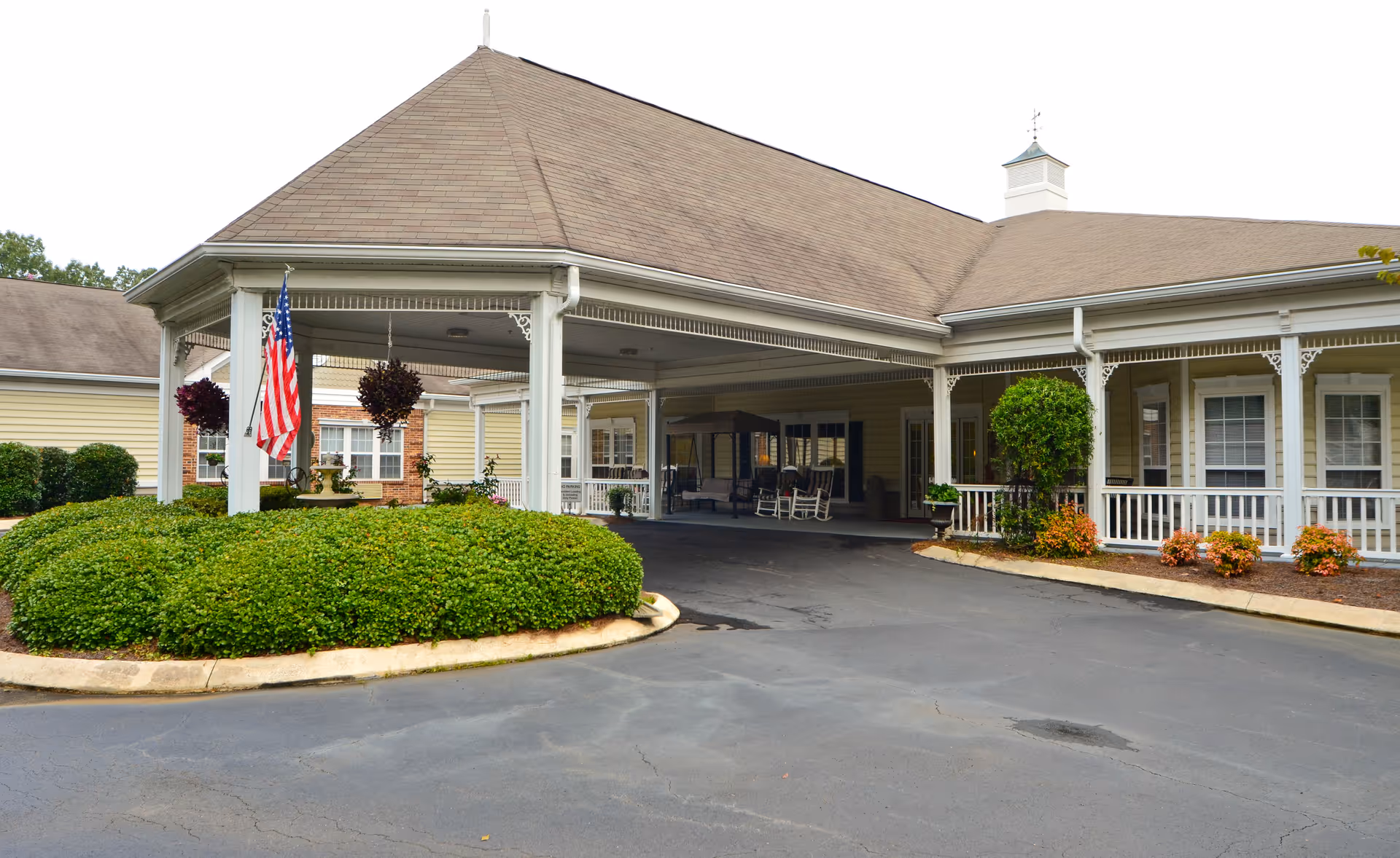 Exterior view of Lancaster Grove Senior Living facility showing a covered entrance with white pillars, an American flag, hanging flower baskets, and well-maintained bushes and shrubs around the driveway.