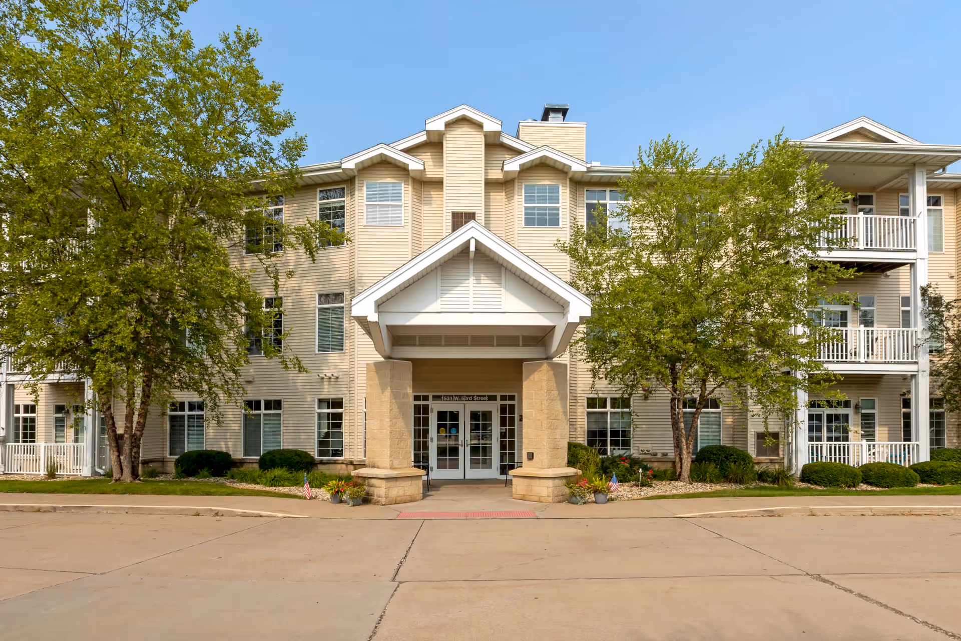 Front entrance of a three-story beige senior living building with trees, balconies, and a covered porte-cochère.