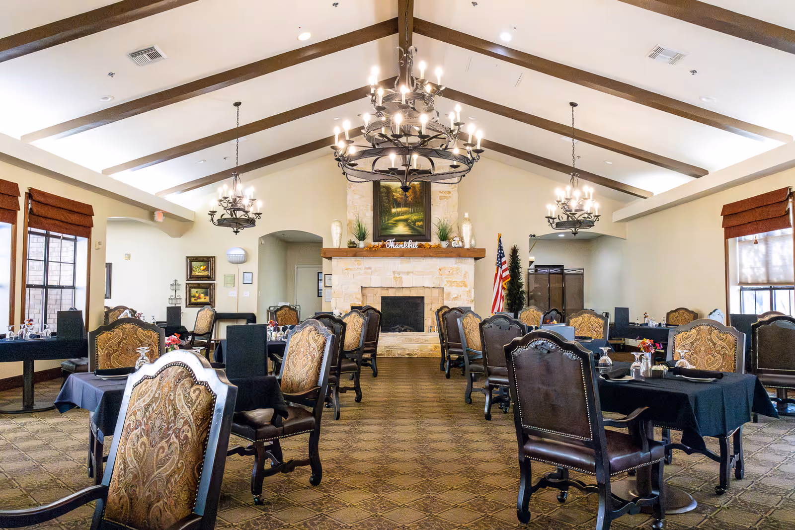 Spacious dining room with multiple set tables and ornate chairs, chandeliers, and a stone fireplace.