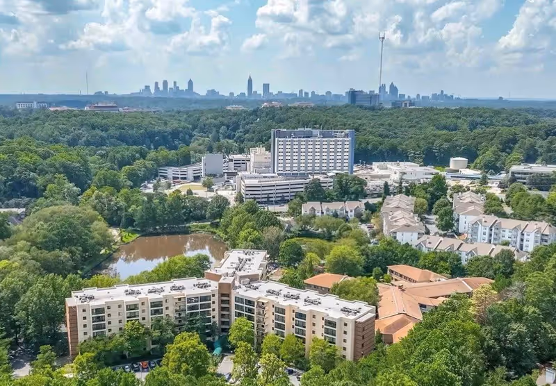 Aerial view of Montclair Assisted Living facility surrounded by trees and greenery, with a small pond nearby and a city skyline visible in the distance under a partly cloudy sky.
