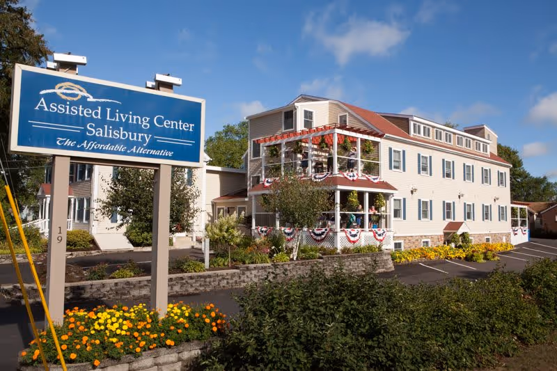Exterior front view of the Assisted Living Center - Salisbury with a large blue sign, decorated porch, and landscaped flowerbeds.