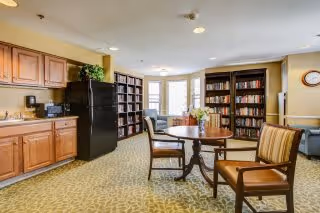 A cozy common area in a senior living facility featuring a small round wooden table with two cushioned chairs. The room includes wooden cabinetry with a black refrigerator, bookshelves filled with books, and large windows allowing natural light to brighten the space. The carpet has a subtle pattern, and the walls are painted in a warm beige tone.