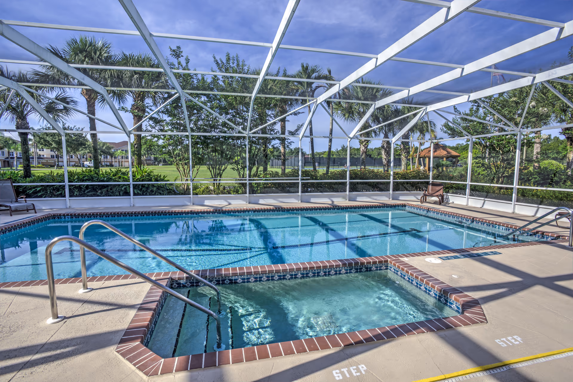 An outdoor swimming pool and a smaller hot tub enclosed by a white screened structure. There are palm trees and greenery visible outside the enclosure under a clear blue sky. Poolside chairs are placed around the pool area.