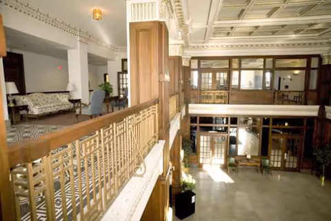 Two-story sunlit senior living lobby with wood railings, seating areas, and glass doors.
