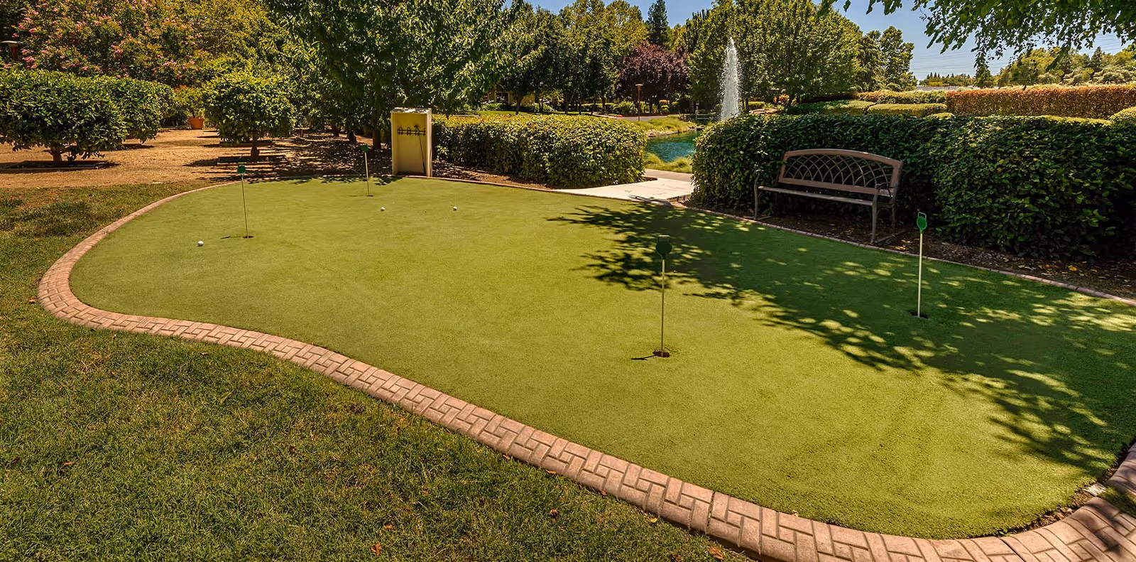 Outdoor putting green with four golf holes and golf balls, surrounded by green grass, bushes, trees, a bench, and a water fountain in the background under a clear sky.