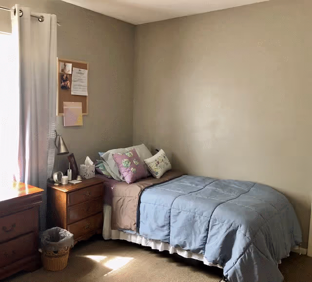 Small furnished bedroom with a twin bed against a beige wall, a nightstand with lamp and personal items, and a curtained window.