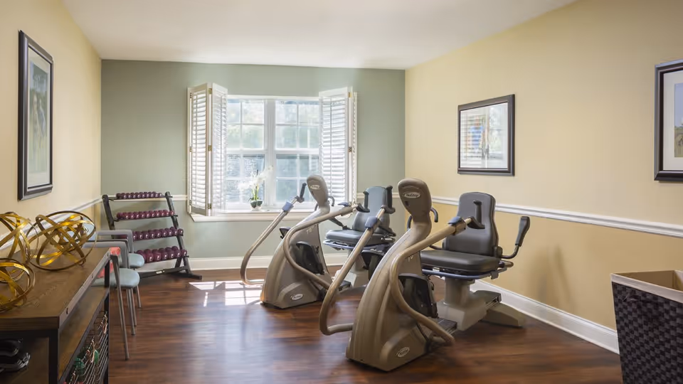 A small exercise room with two recumbent stationary bikes facing a window with white shutters. There is a rack of purple dumbbells in the corner, a wooden console table with decorative golden spheres, and two chairs along the left wall. The room has light yellow and green walls with framed pictures and wood flooring.