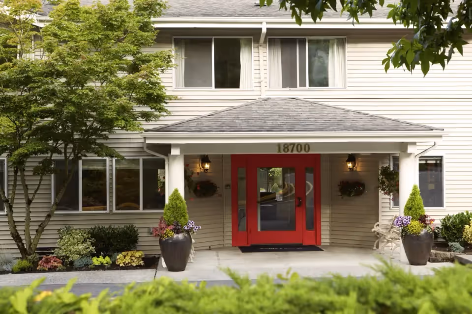 Entrance of a building with beige siding and a gray shingled roof. The entrance features a red double door under a small covered porch with the number 18700 above it. There are two large planters with flowers and greenery on either side of the door, wall-mounted lantern lights, and a bench to the right. Trees and shrubs surround the entrance area.