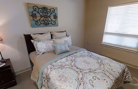 A neatly made bed with patterned bedding in soft blue, beige, and white tones in a bedroom. There is a wooden nightstand with a lamp on the left side of the bed. A decorative wall art piece with a wooden background and black wrought iron design hangs above the bed. A window with white blinds is on the right wall.