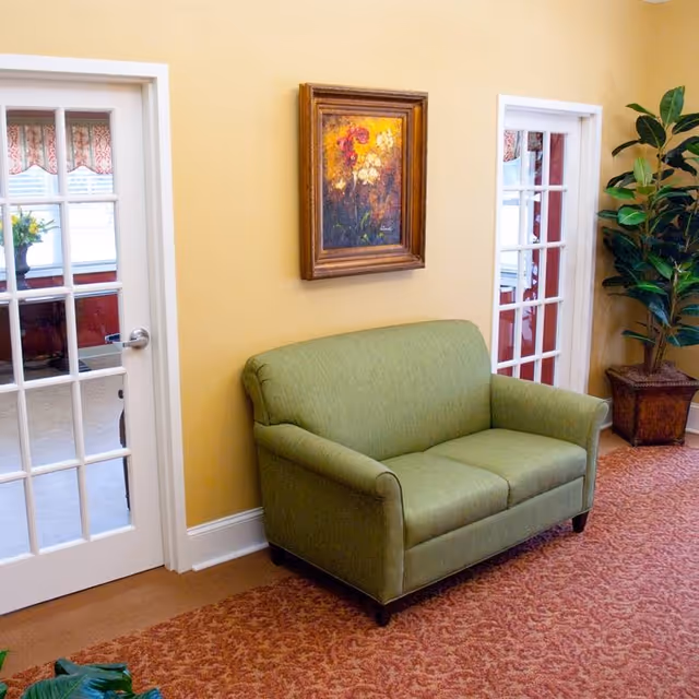 Green two-seat sofa against a yellow wall flanked by two glass-paned doors, with a framed painting above and a potted plant nearby.