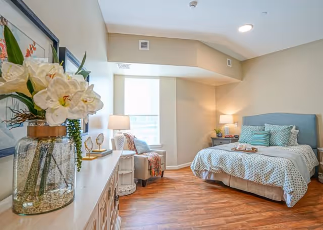 Sunlit furnished bedroom with a bed, bedside tables, an armchair by a window, and a dresser topped with a vase of flowers.
