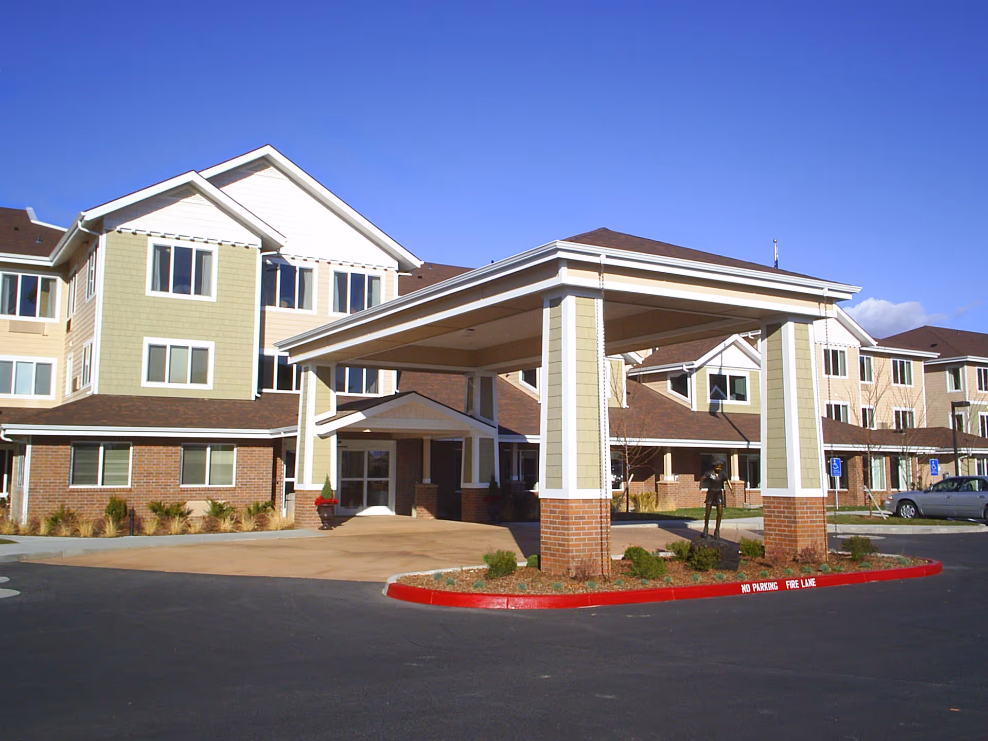 Exterior view of a multi-story senior living facility with a covered entrance driveway, landscaped areas, and clear blue sky.