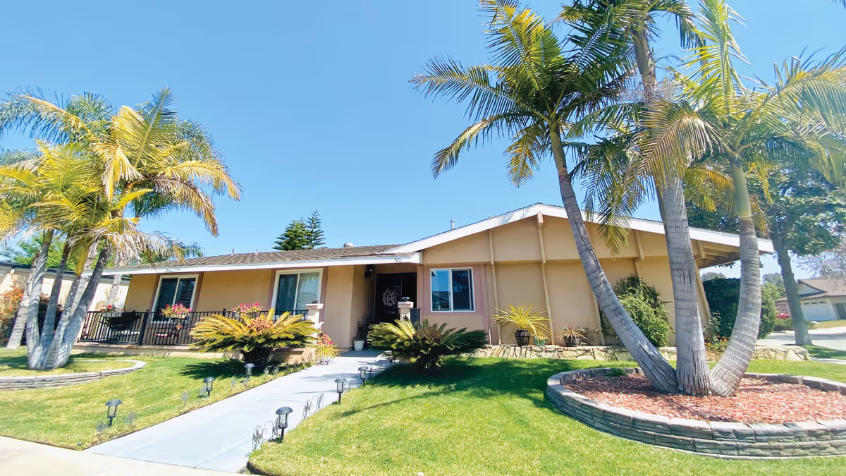 Single-story residential building with a beige exterior, surrounded by a well-maintained lawn and several palm trees under a clear blue sky. A concrete pathway leads to the front door, and there are small garden lights lining the path.