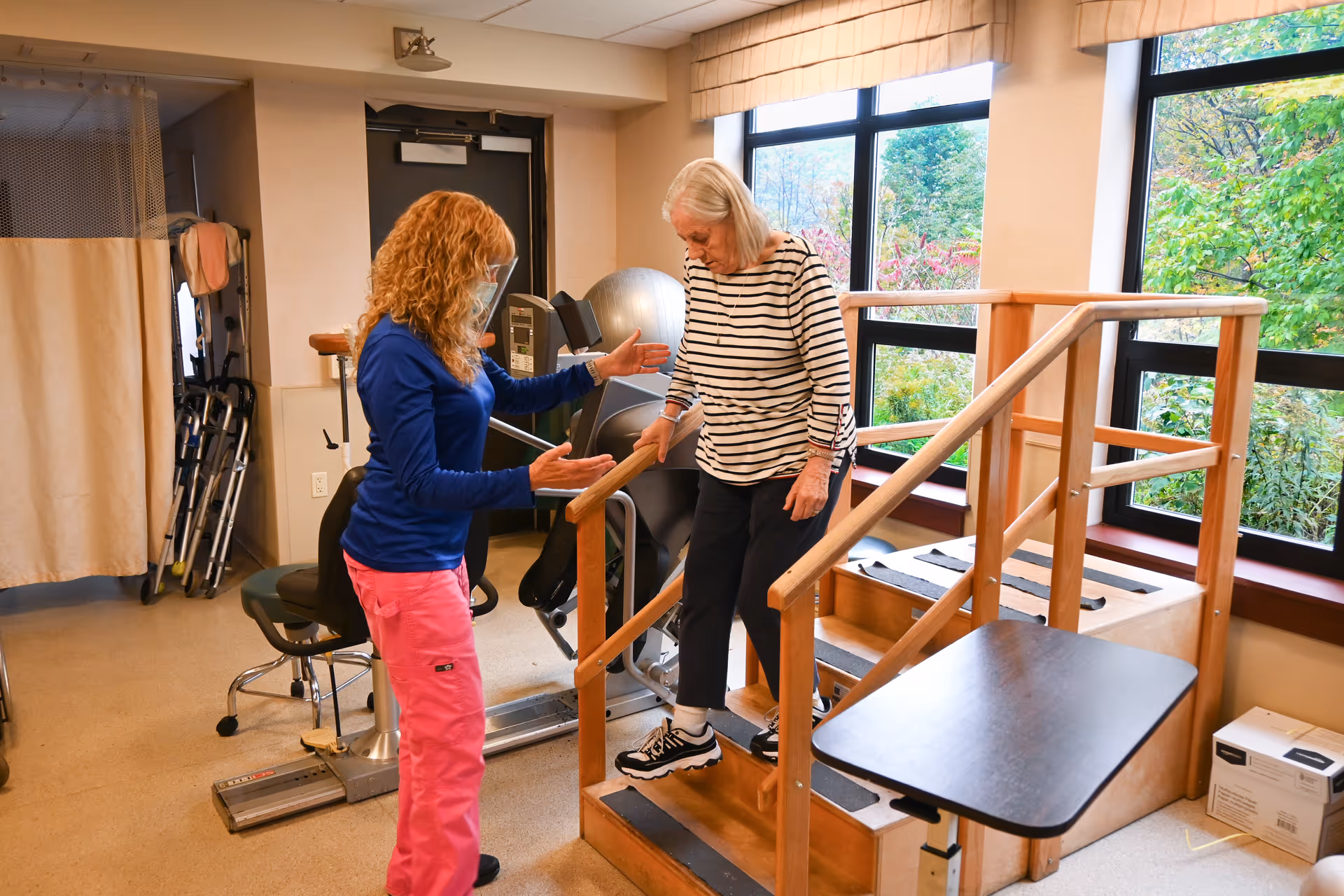A physical therapist assists an elderly woman as she practices walking down wooden stairs with handrails inside a rehabilitation room with exercise equipment and large windows showing greenery outside.
