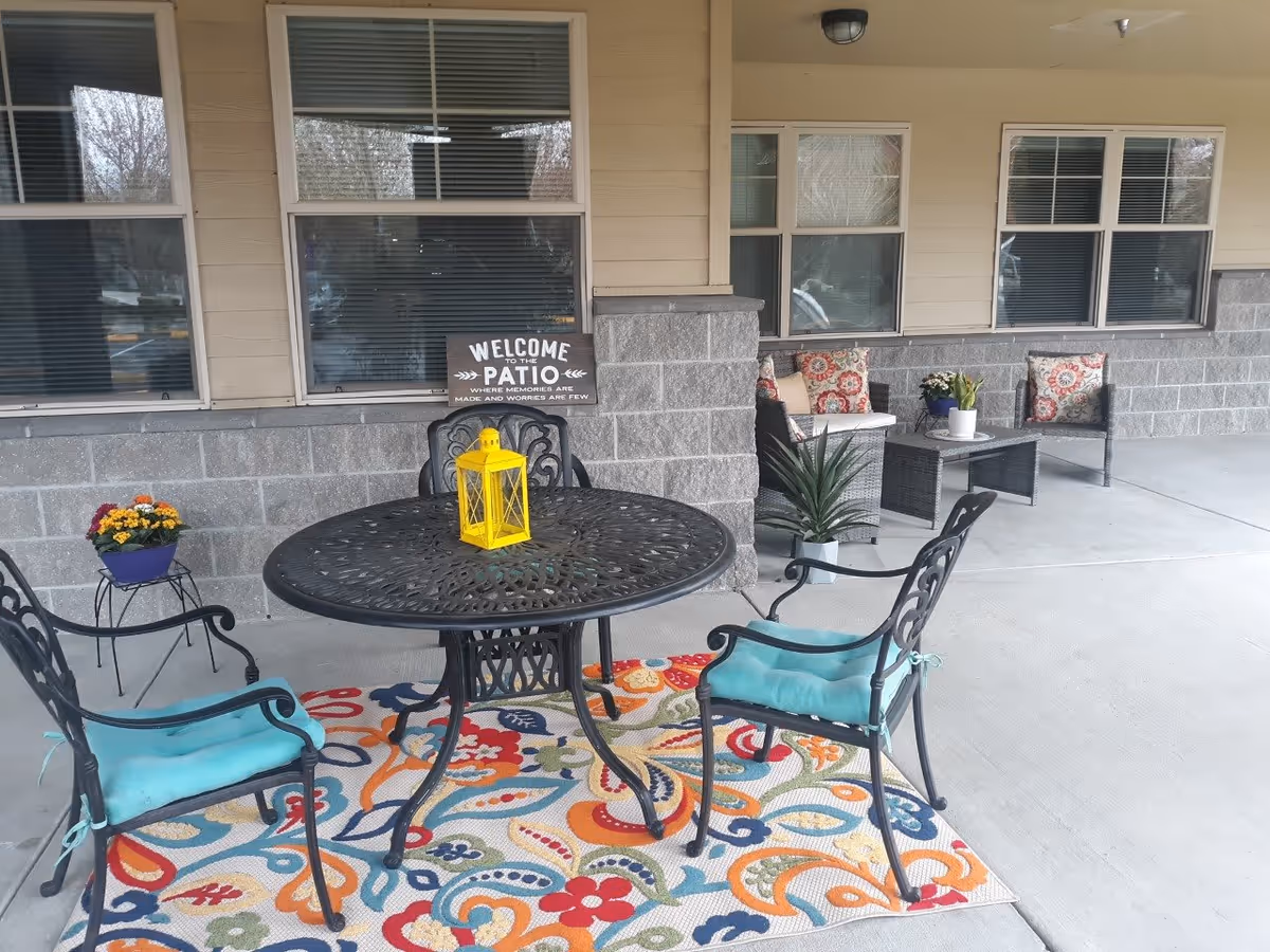 Outdoor patio area with a round black metal table and four chairs with blue cushions on a colorful patterned rug. A yellow lantern sits on the table. In the background, there are two cushioned chairs with floral pillows and a small table with potted plants. A sign on the wall reads 'Welcome to the Patio where memories are made and worries are few.'