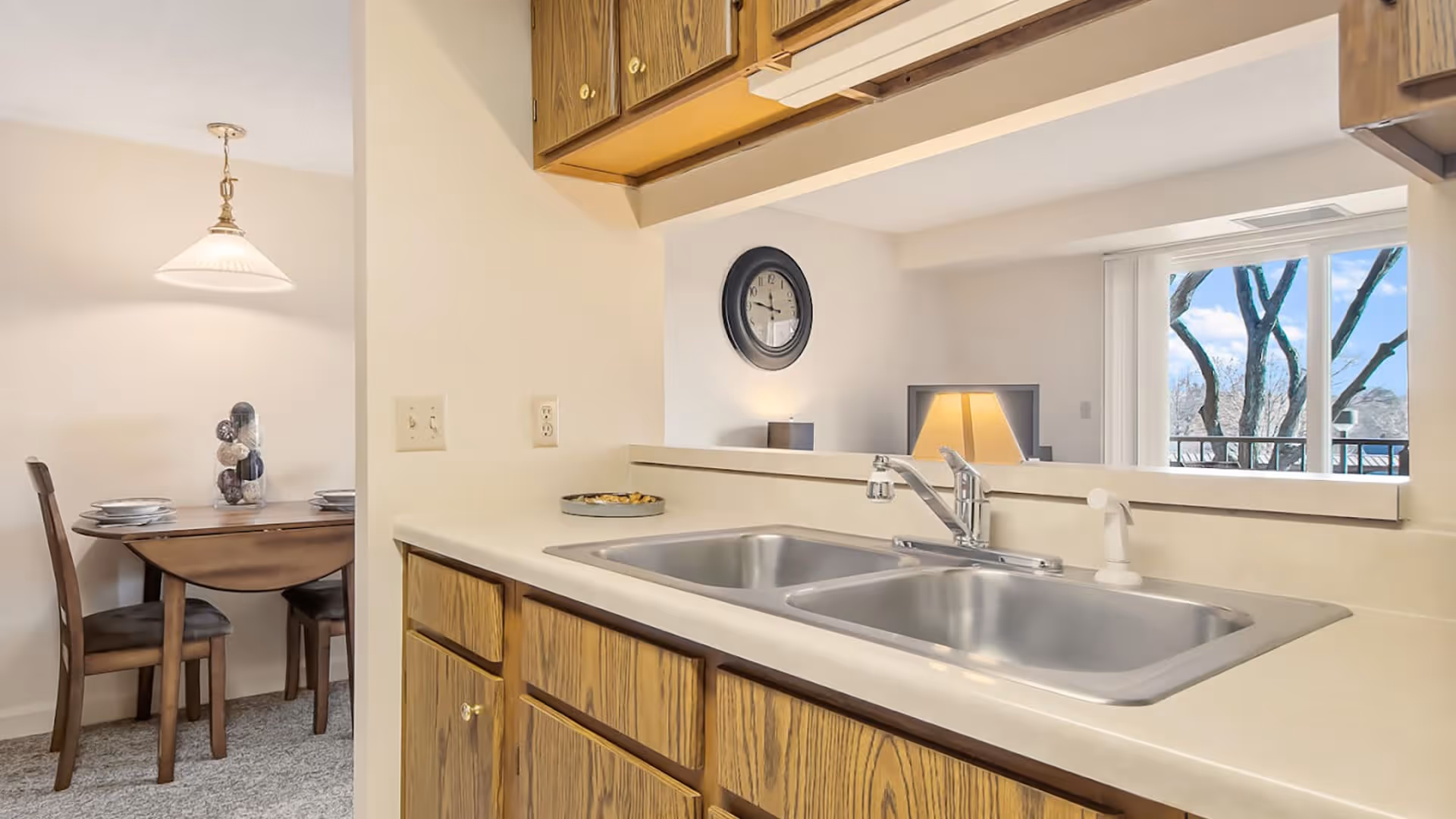 Interior view of a kitchen area with a double stainless steel sink and wooden cabinets. In the background, there is a dining area with a wooden table set for four and a hanging light fixture. A living room area with a clock on the wall, a lamp, and a sliding glass door showing trees outside is also visible.