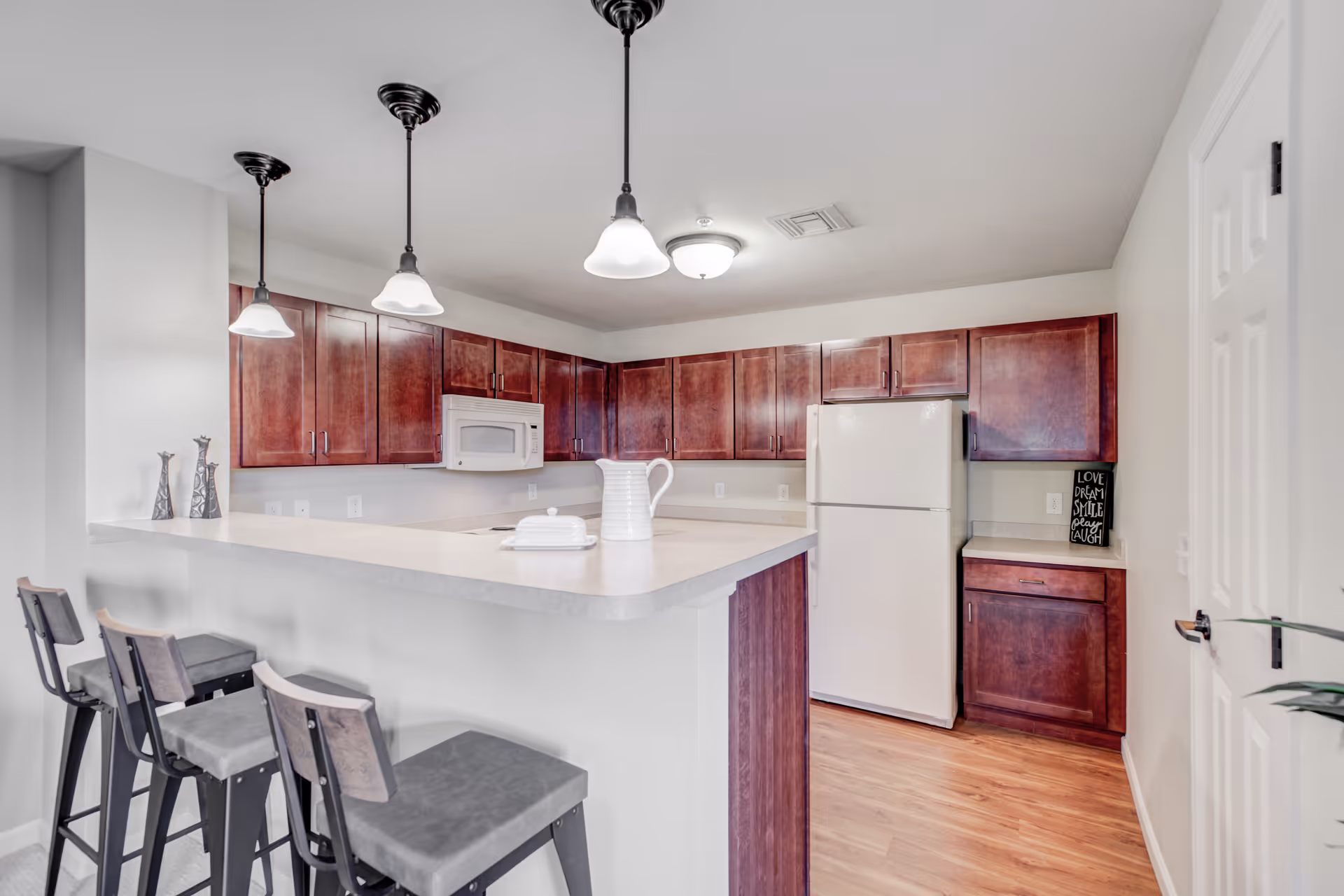 A modern kitchen with dark wood cabinets, a white refrigerator, a white microwave, and a light-colored countertop with a raised bar area. Three pendant lights hang above the bar, which has three high chairs. The floor is wooden, and there is a small decorative sign on the counter that reads 'Love Dream Smile Laugh.'
