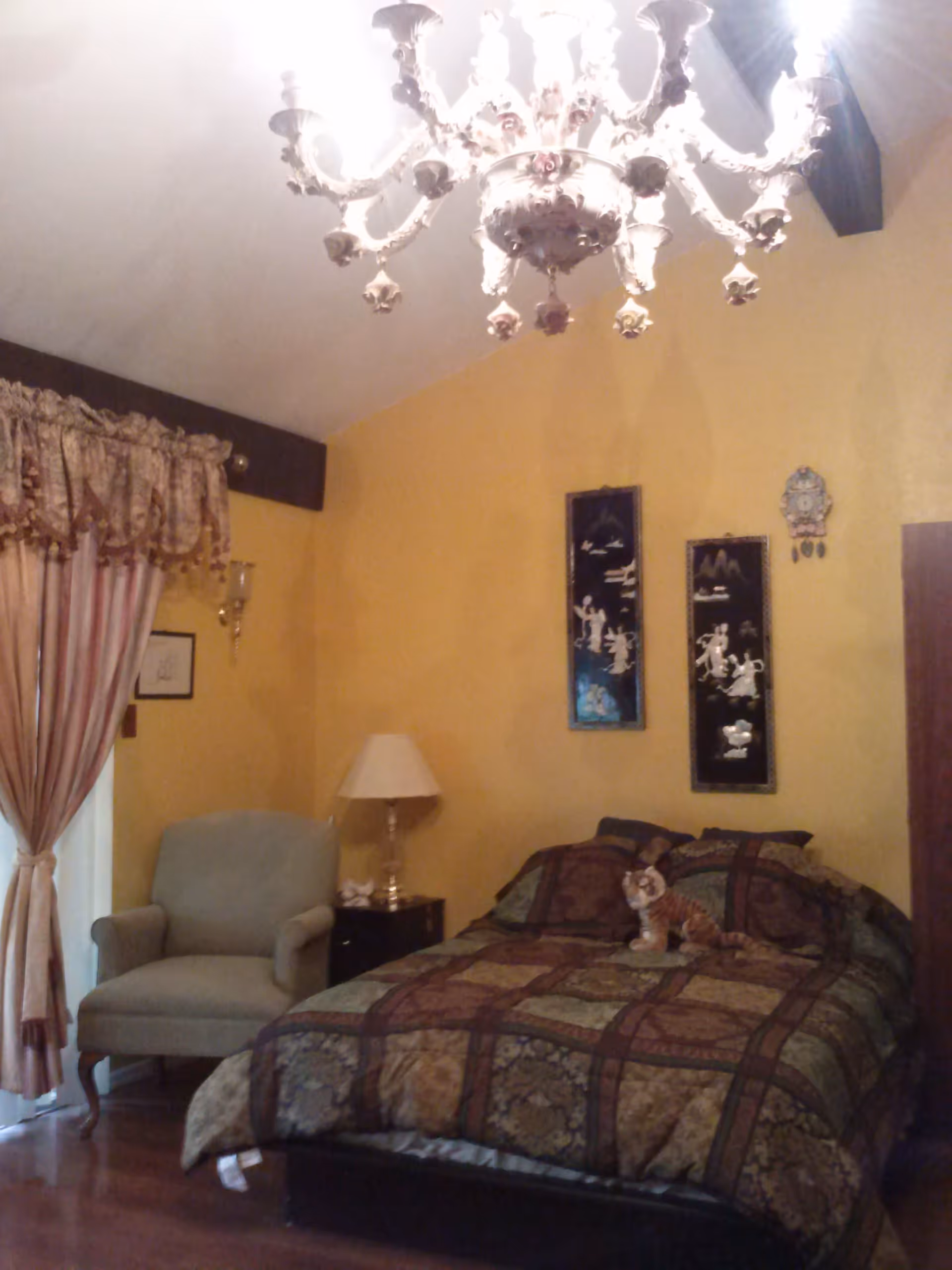 A cozy bedroom with yellow walls, a bed covered with a patterned quilt and a stuffed tiger toy on it, a beige armchair near a window with pink and gold curtains, a nightstand with a lamp, and two framed artworks hanging on the wall. A decorative chandelier hangs from the ceiling.