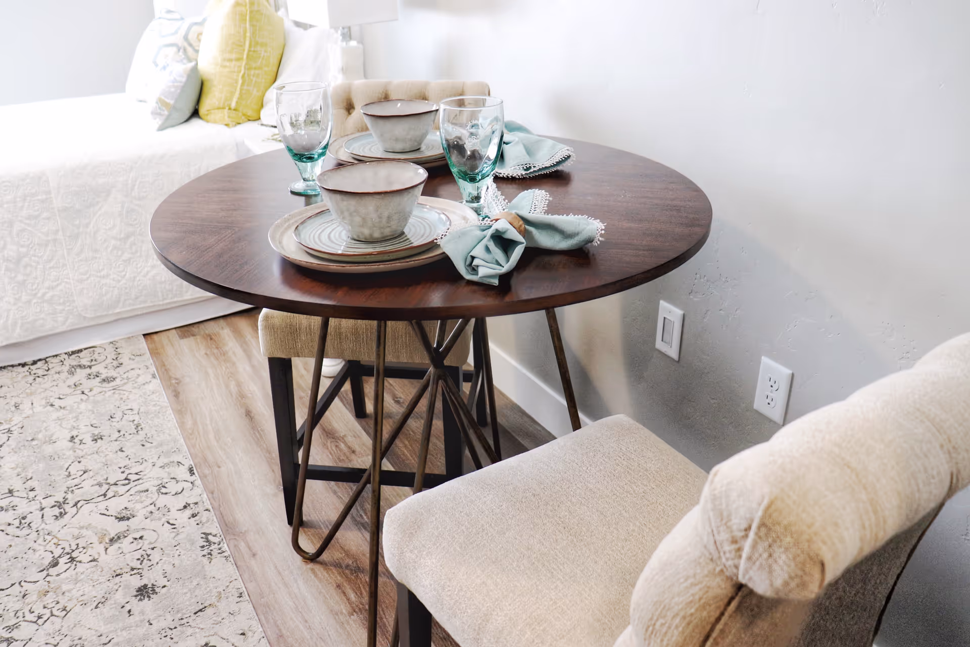A small round wooden dining table set for two with ceramic bowls, plates, glasses, and cloth napkins. Two beige upholstered chairs are positioned around the table. The table is placed next to a bed with white bedding and decorative pillows, and the floor has wood and patterned rug sections.