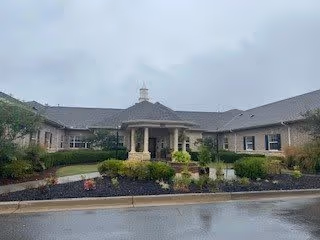 Front exterior view of Peachtree Villa at Suwanee, a single-story building with a covered entrance supported by columns, surrounded by landscaped greenery and a driveway in front.