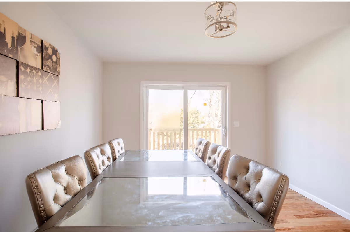 A bright dining room with a long glass-top table surrounded by eight tufted chairs. There is a modern light fixture on the ceiling and a sliding glass door at the far end leading to an outdoor area. A multi-panel wall art piece is displayed on the left wall.