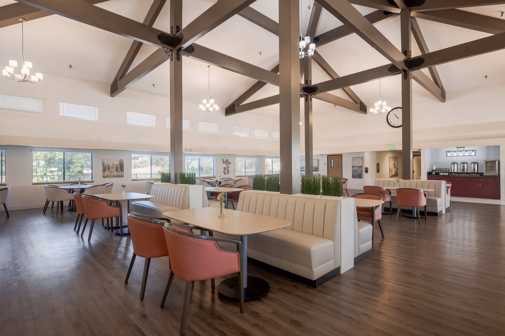 A spacious dining room with high vaulted ceilings featuring exposed wooden beams and multiple chandeliers. The room has several tables with white cushioned booth seating and pink chairs. Large windows line the walls, allowing natural light to fill the space. In the background, there is a counter area with coffee dispensers and other amenities.