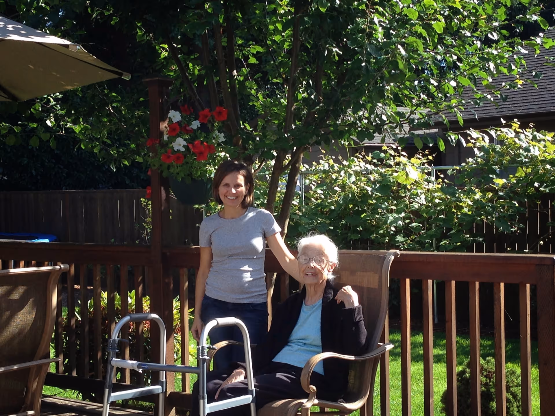 An elderly woman sitting in a chair on a wooden deck outdoors with a younger woman standing beside her, smiling. There is a walker in front of the elderly woman, hanging flower baskets, green trees, and a wooden fence in the background.