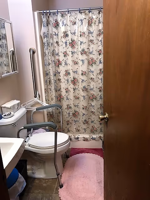 Small bathroom with a toilet equipped with safety rails, a floral shower curtain, a pink bath mat, a sink, and a wooden door partially open.