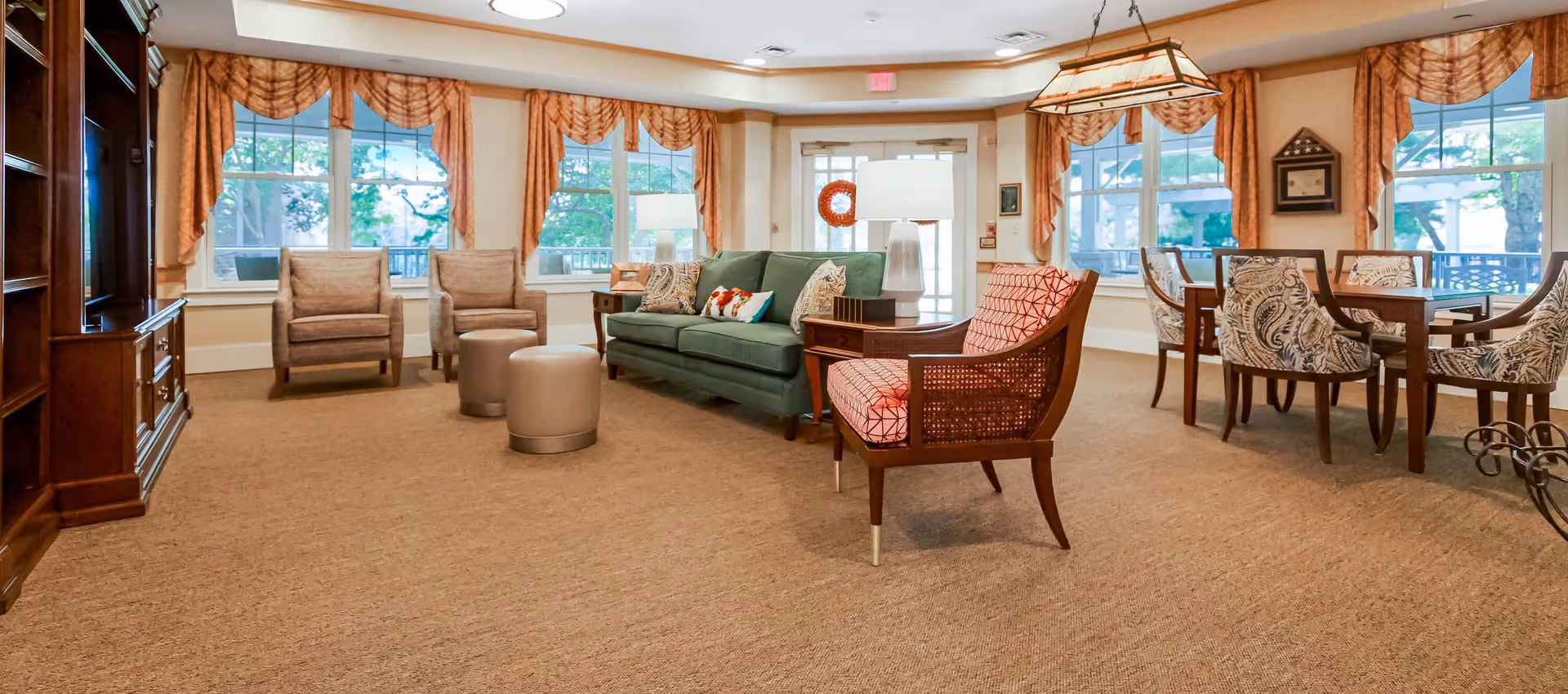 Communal living room with upholstered chairs, a green sofa, round ottomans, and a dining table by large windows.
