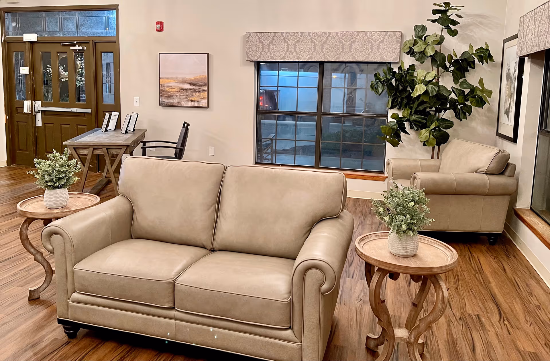 A cozy communal living area with beige leather sofas, wooden side tables topped with potted plants, a desk by the entrance, and a large window.