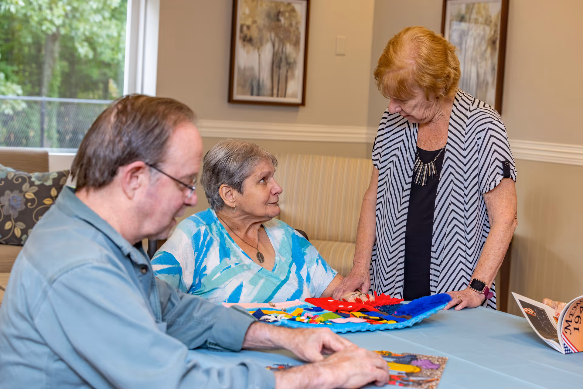 Three elderly individuals engaged in an activity at a table inside a well-lit room. One woman is seated wearing a blue and white patterned shirt, looking up at another standing woman with red hair wearing a black and white striped cardigan. A man with glasses and a green shirt is focused on a puzzle or similar activity on the table. The room has beige walls, framed artwork, and a window showing greenery outside.