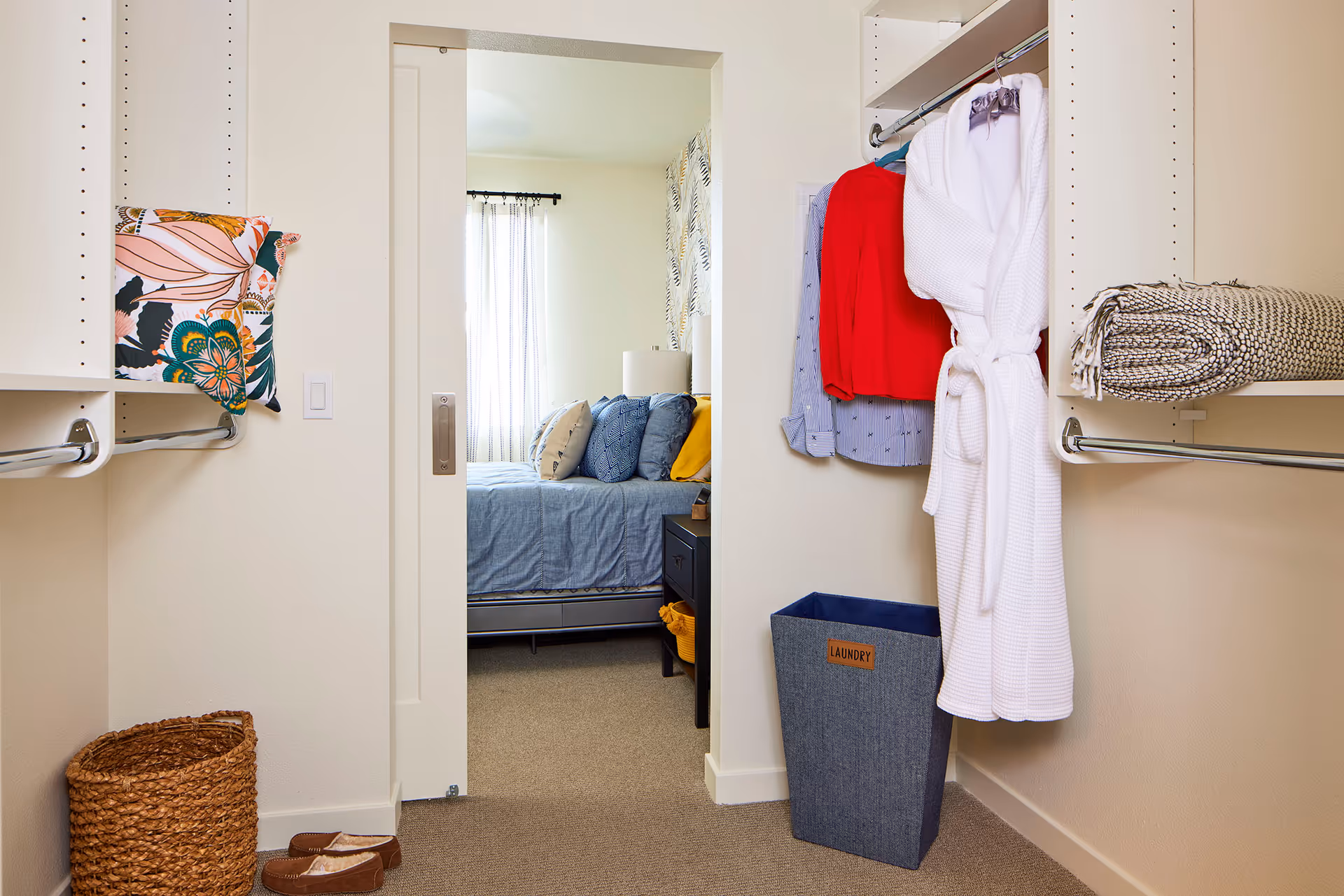 View from a walk-in closet into a bedroom with hanging clothes, a laundry bin, a woven basket, and a bed with pillows.