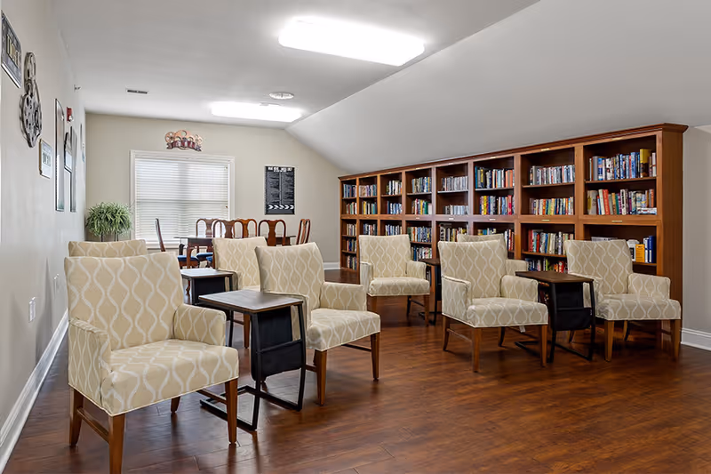 A cozy reading and seating area in a senior living facility with six patterned armchairs arranged in two rows, each paired with a small wooden side table. Behind the chairs is a large wooden bookshelf filled with books. In the background, there is a dining table with chairs near a window with blinds, and a potted plant on the left side. The room has wooden flooring and soft overhead lighting.