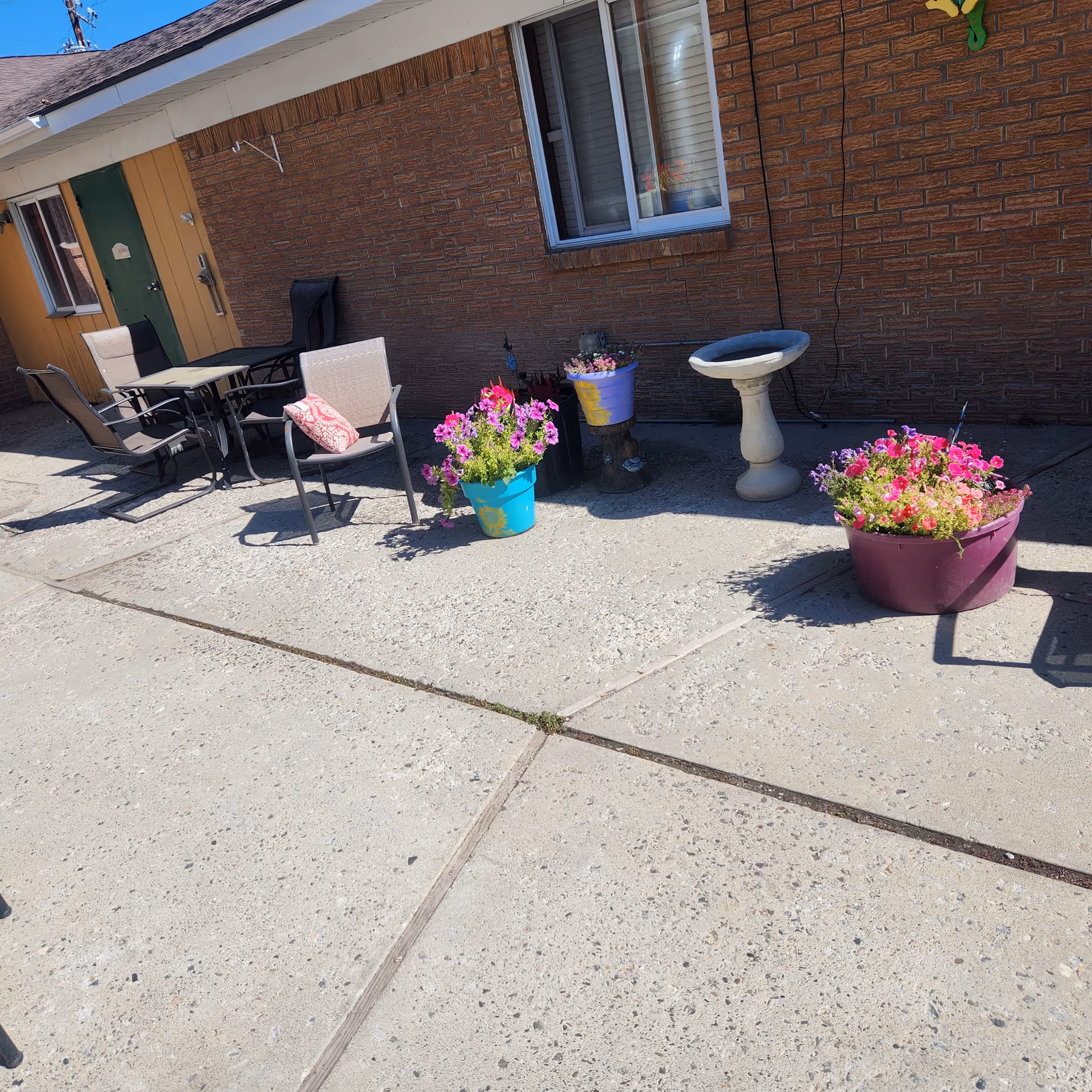 Outdoor patio area with several chairs and a table arranged near a brick wall. There are colorful flower pots with blooming flowers and a birdbath placed along the wall. The ground is concrete, and the scene is brightly lit by sunlight.