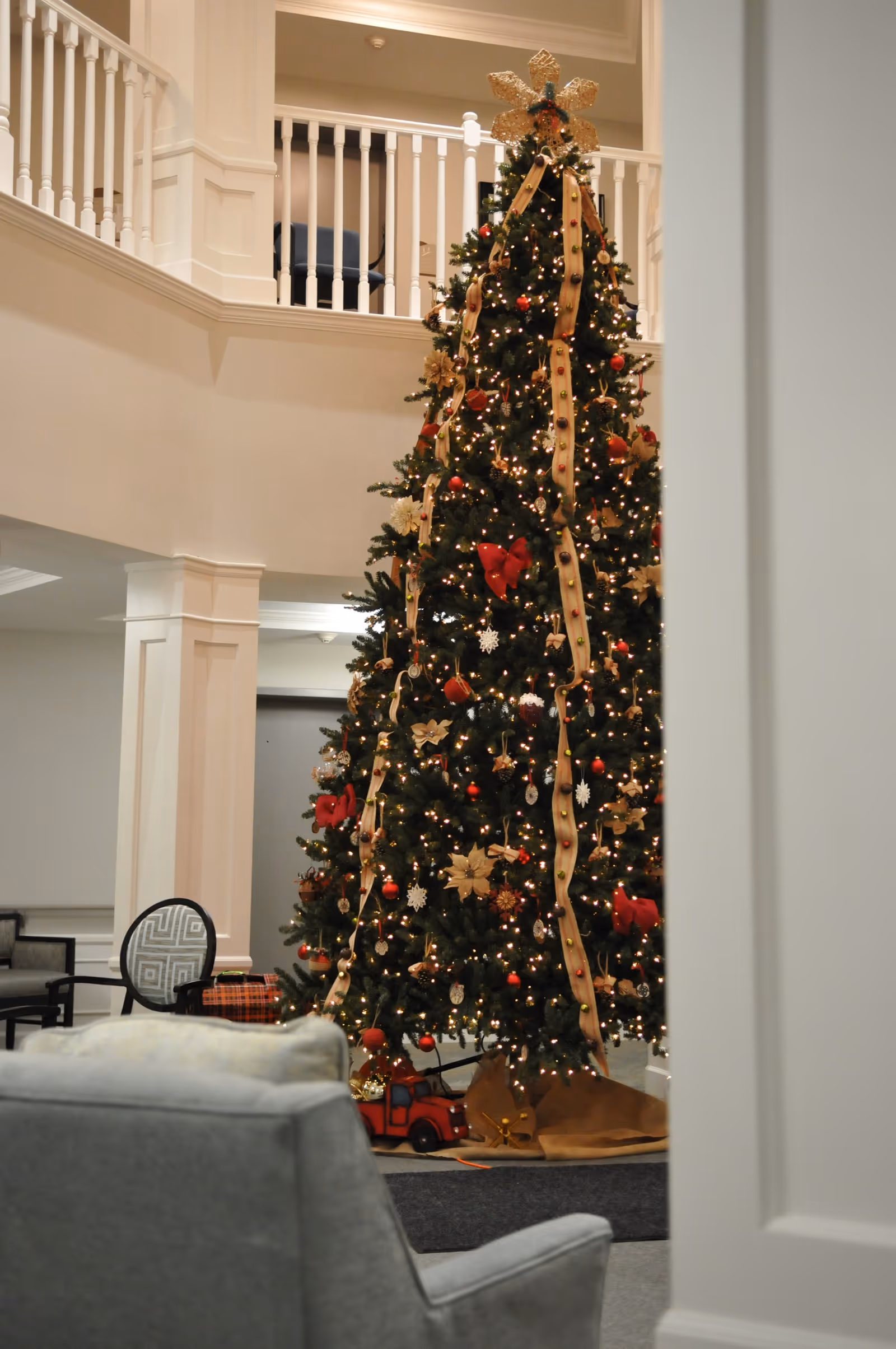 A tall, decorated Christmas tree with lights, ribbons, and ornaments stands in a spacious, well-lit interior area with white walls and railings. There are chairs and a small table nearby, and the setting appears to be a common area or lounge in a facility.