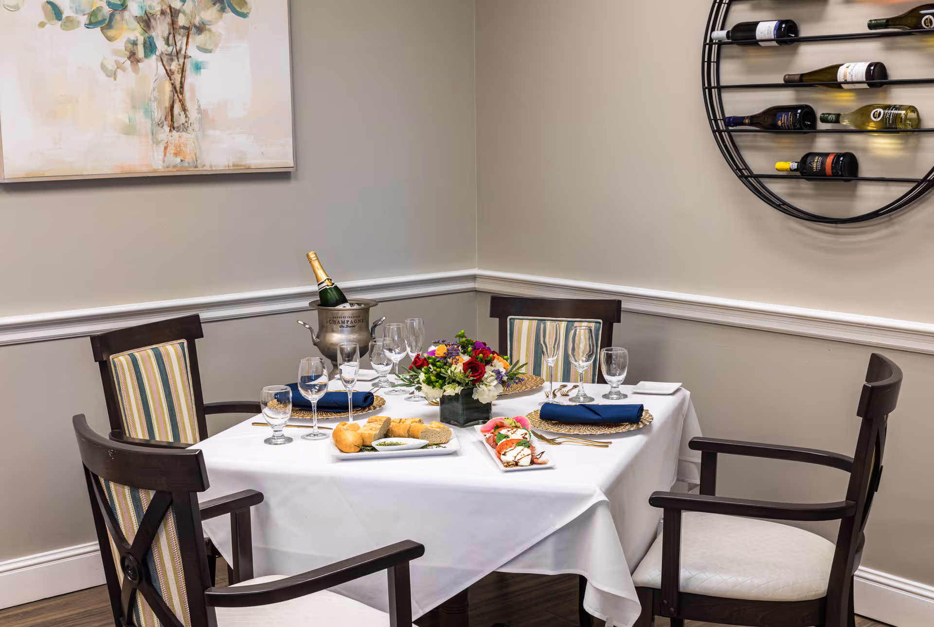 A small dining table set for four with a white tablecloth, blue napkins, wine glasses, water glasses, and gold utensils. The table has a centerpiece of colorful flowers, a bottle of champagne in an ice bucket, a plate of bread, and a plate of sliced tomatoes with mozzarella and basil. The chairs have dark wood frames with striped cushions. On the wall, there is a round wine rack holding several bottles and a painting with abstract floral art.