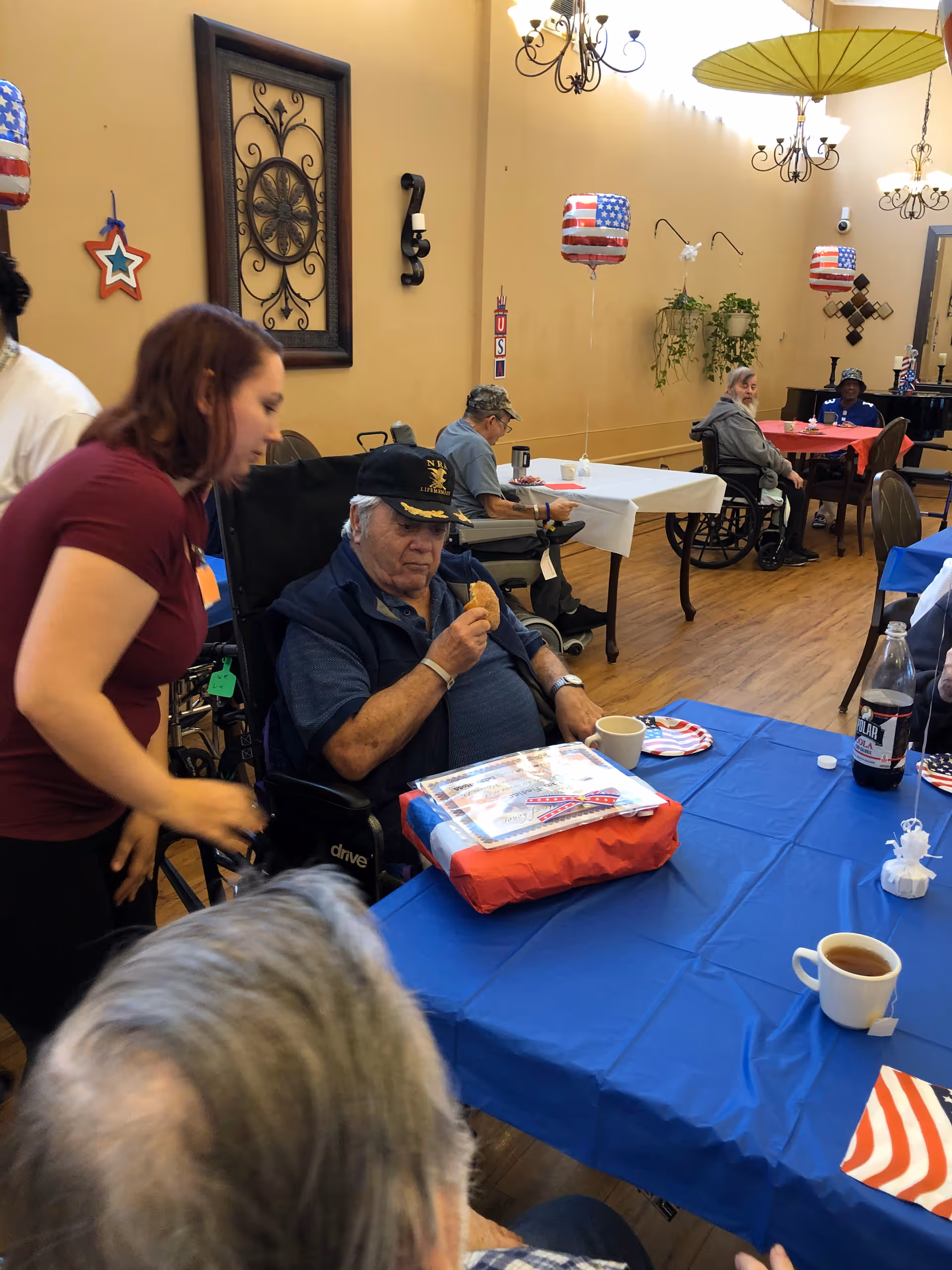 A group of elderly individuals, some in wheelchairs, seated around tables covered with red, white, and blue tablecloths in a decorated dining area. A woman is leaning over to interact with one of the men in a wheelchair who is holding a cookie. The room has patriotic decorations including balloons with American flag designs and wall hangings.
