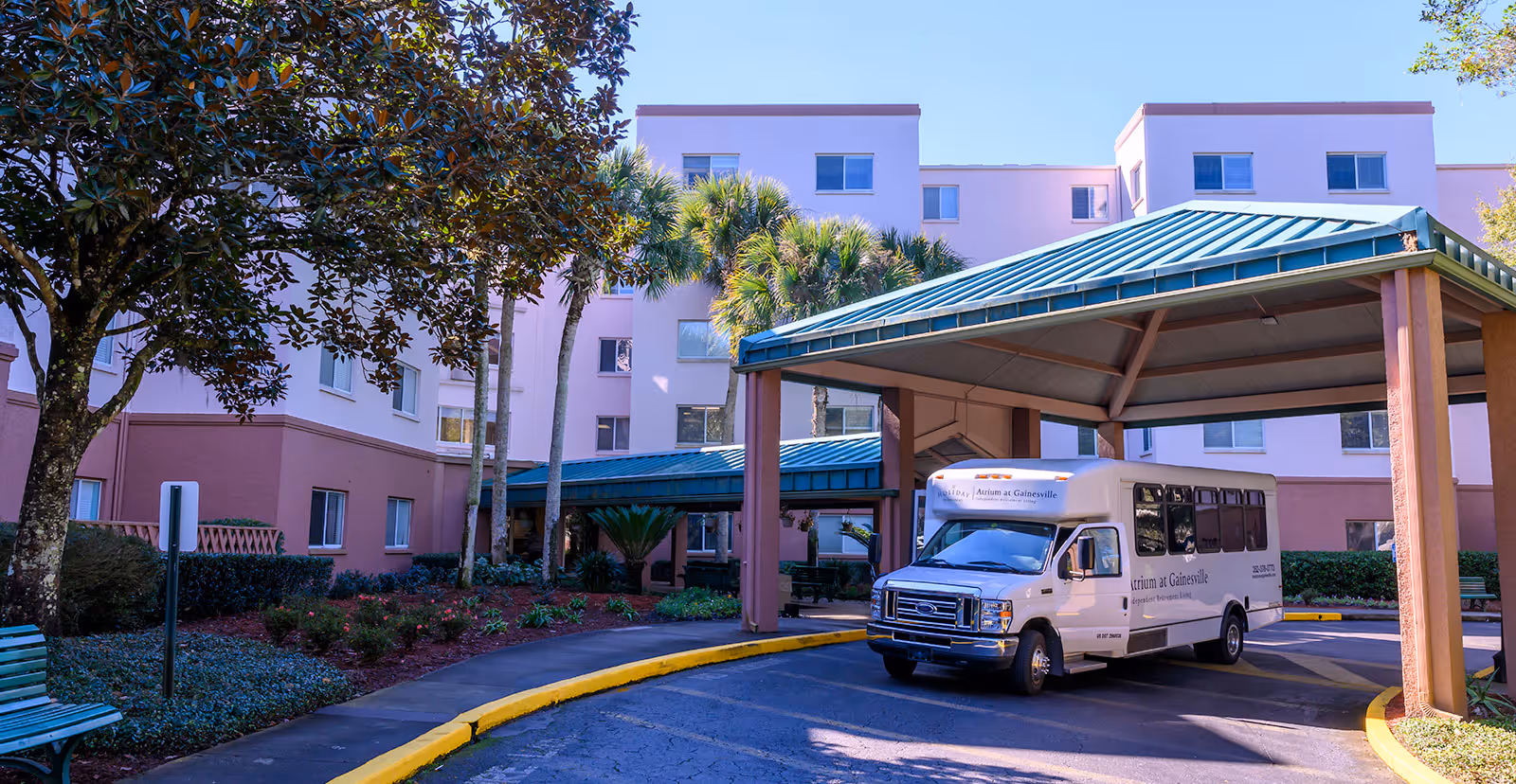 Exterior view of Atrium at Gainesville senior living facility showing a covered entrance with a white shuttle bus parked underneath. The building is light pink with multiple windows and surrounded by palm trees and landscaping.